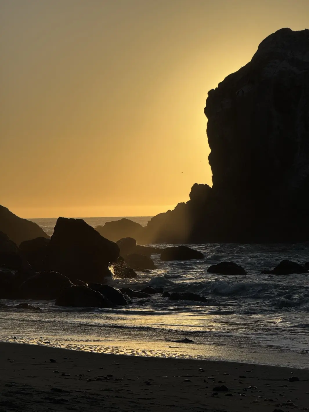 Evening sun bursting past a large sea stack, light spilling across the water.